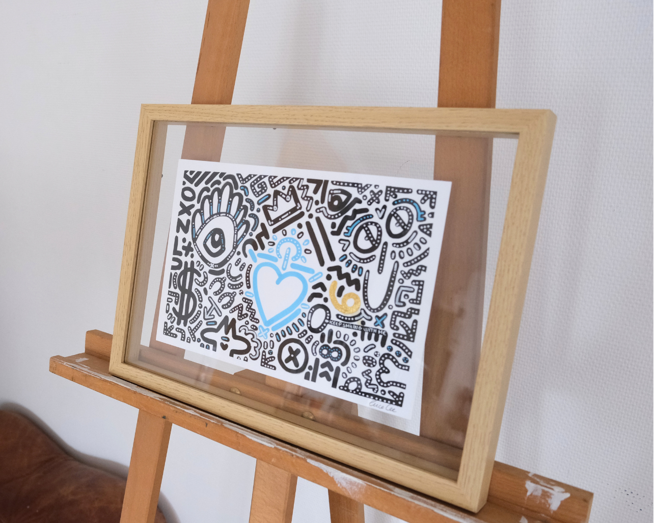 Œuvre originale Keep Smiling With Me n°14 présentée dans son cadre, dessin noir avec touche de bleu et feuille d’or.